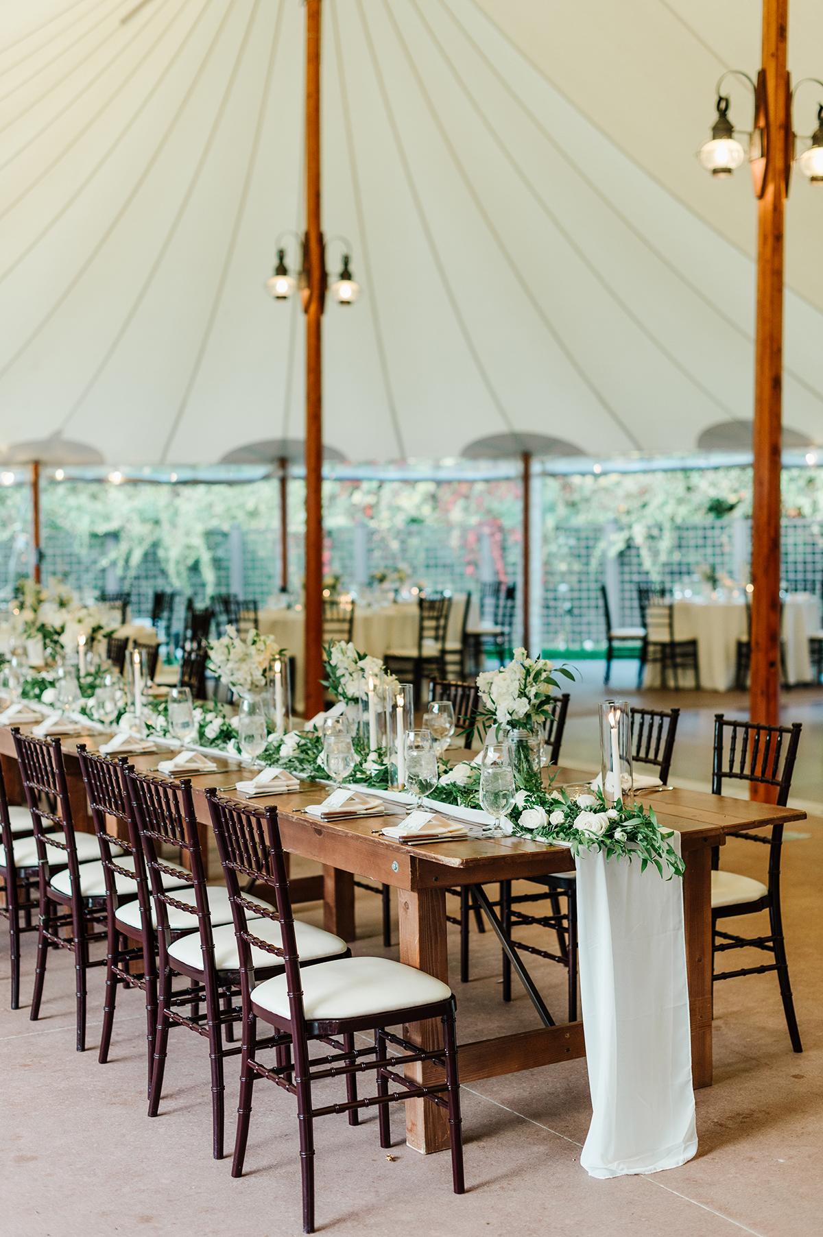 Elegant banquet table set with greenery and white flowers under a tent.