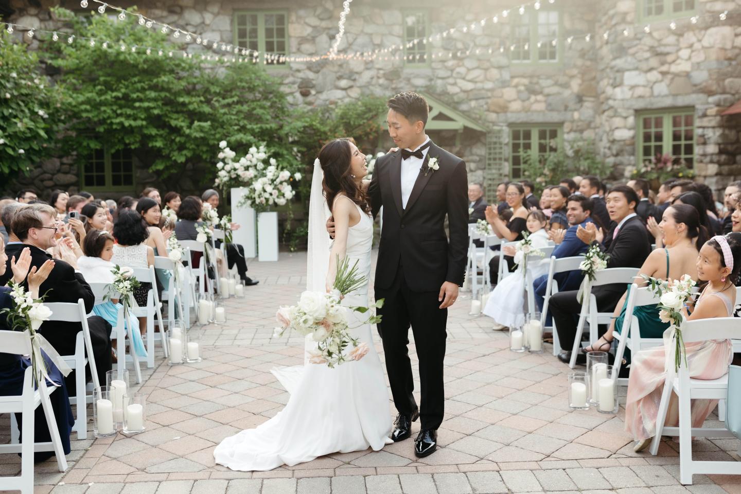 Bride and groom smile, walking down aisle surrounded by seated guests.